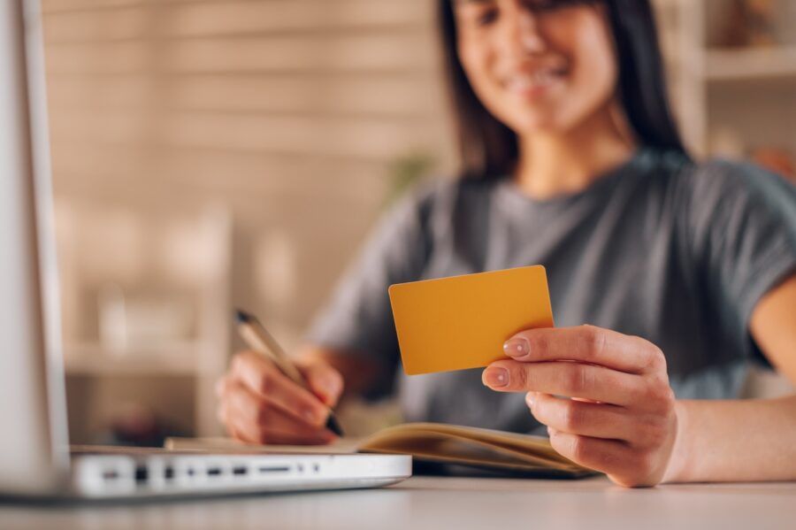 Blurred cropped shot of a dark haired woman holding a credit card while taking notes with a pen in her notebook.