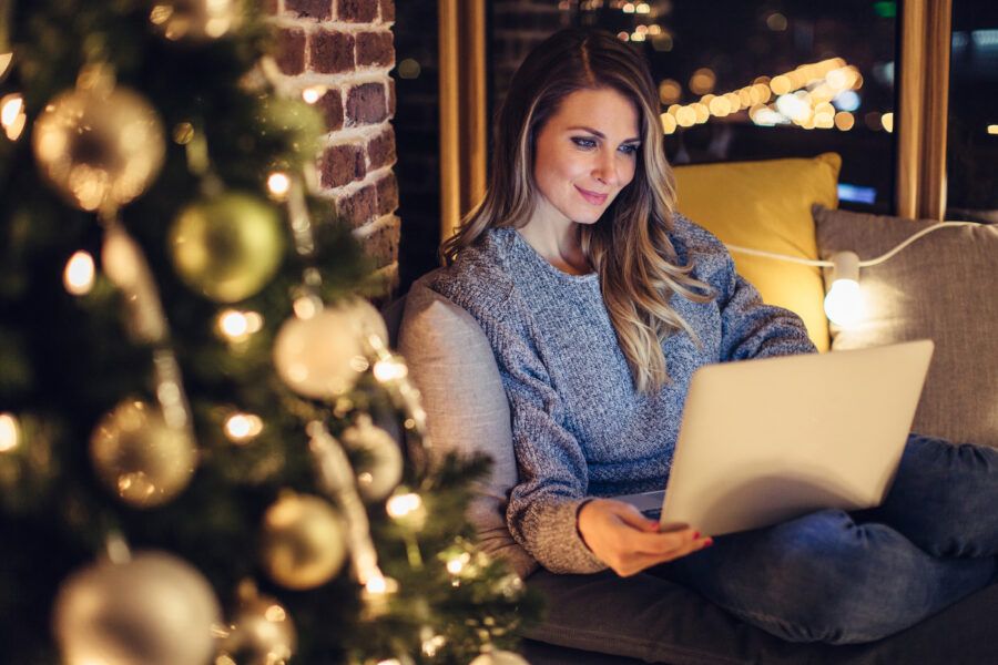 A woman on her couch next to a Christmas tree, shopping online for gifts using a laptop.