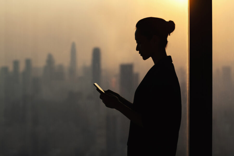 Silhouette of young woman using smartphone next to window with cityscape