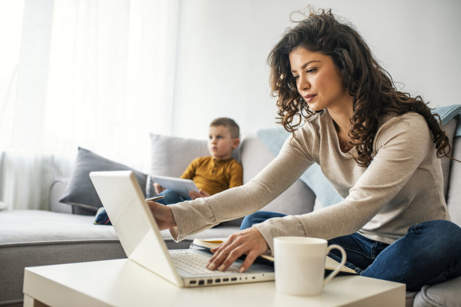 Young mother sitting at table and using laptop at home to read about financial resources for single parents while her child sits on the couch behind her.
