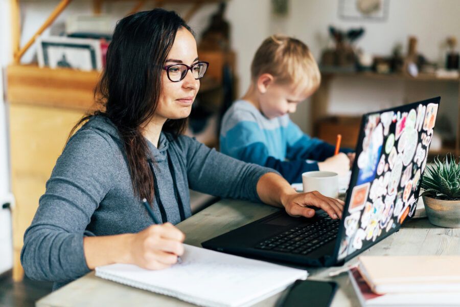 A single parent doing home finances on a laptop while her child draws next to her.