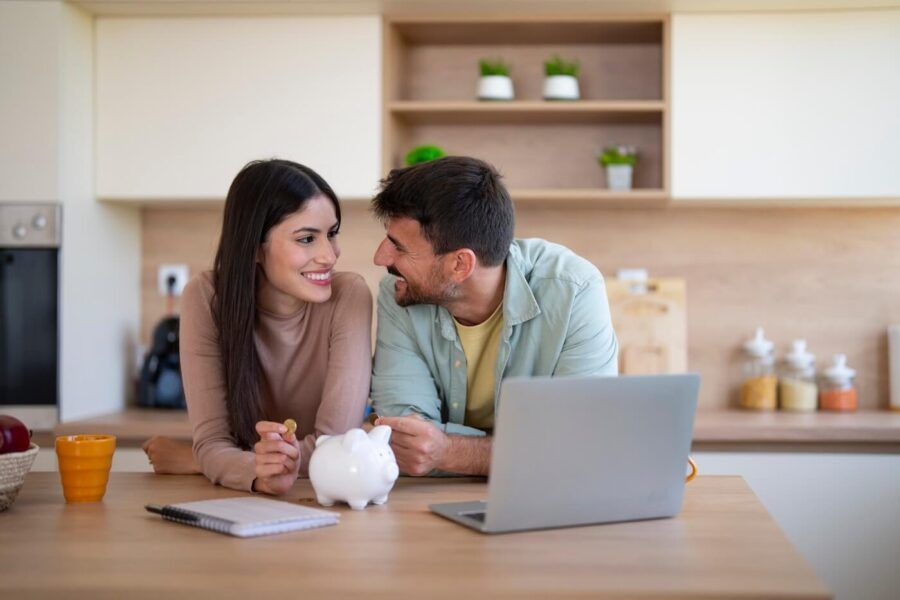Smiling couple sitting at a desk, planning their finances using a laptop and a notepad, with a pink piggy bank beside them