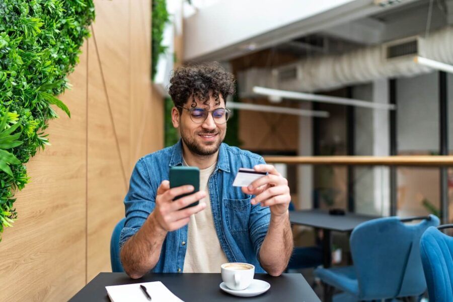 Smiling man seated at a café table holding a smartphone in one hand and a credit card in the other. A cup of coffee and notepad sit on the table, with modern indoor seating, greenery, and natural light visible in the background
