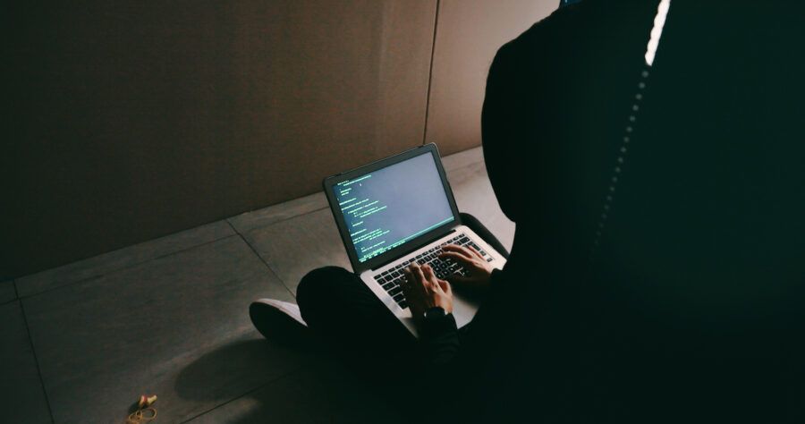 Shot of a social engineer using a laptop in a dark server room, hooded and facing away from the camera.