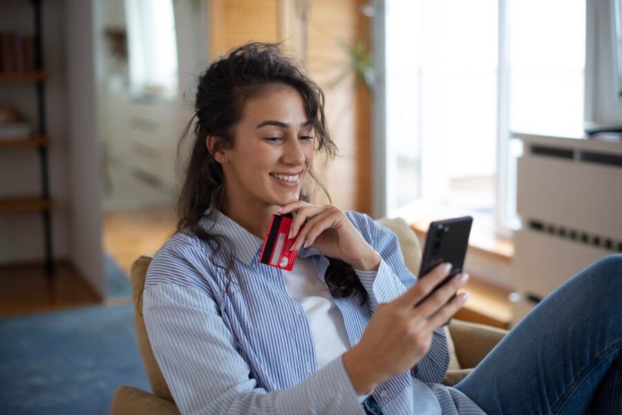 Smiling young woman sitting in a chair while using a smartphone and holding a credit card