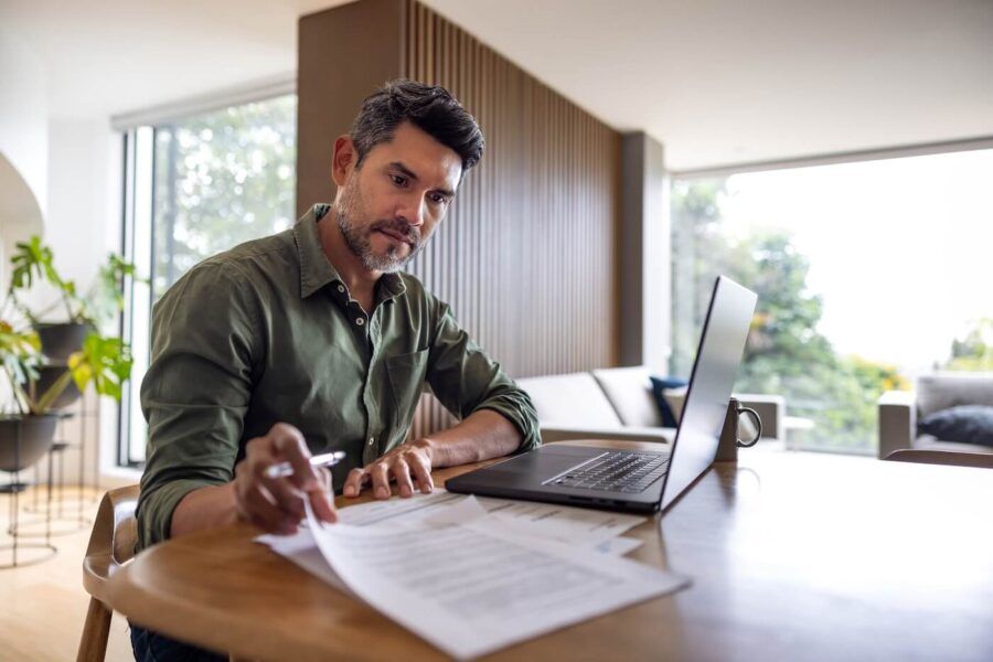 Focused man reviewing printed documents, with an open laptop on a desk