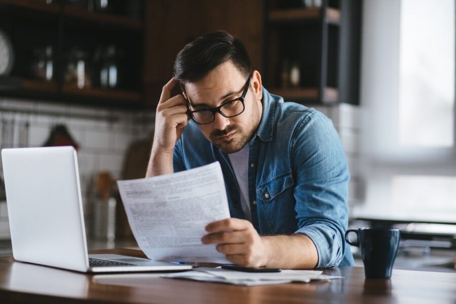 Portrait of a man holding a financial letter while working on his laptop at the kitchen table.