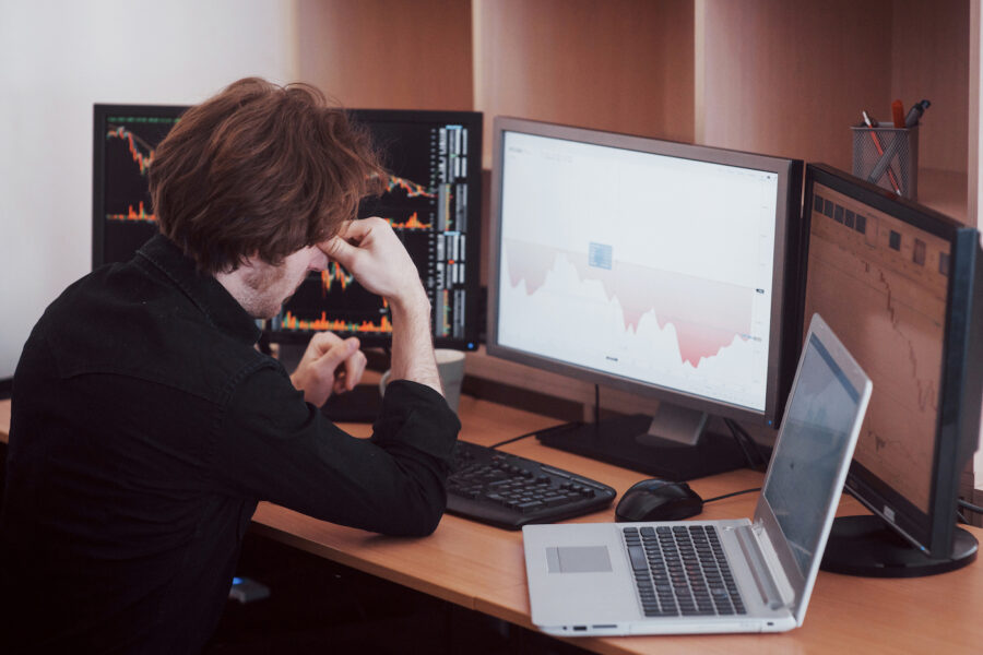 A stressed man staring at his stock performances on his desktop computer, torn between investing and saving in a troubled economy.