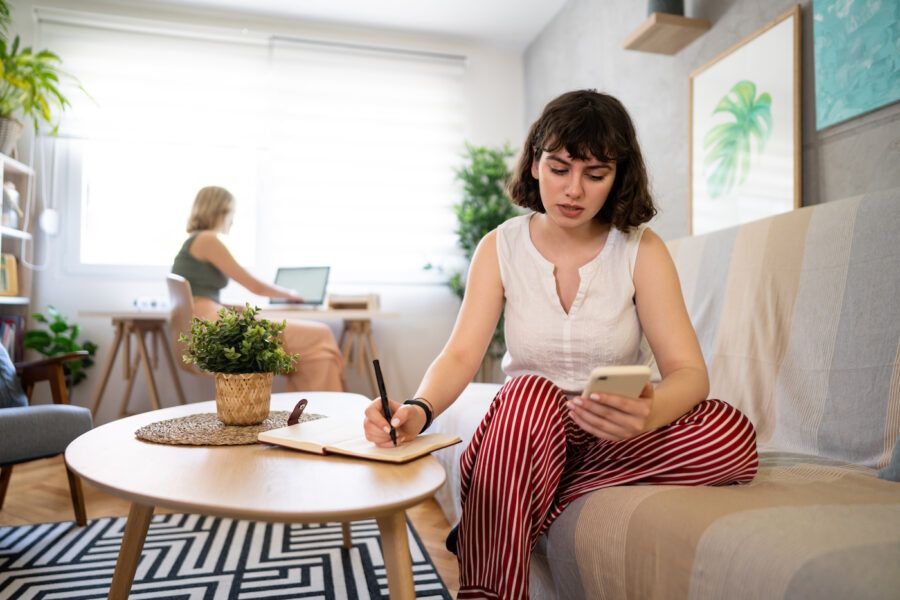 A student sitting on the couch checking her finances on her phone and writing on a notepad on the coffee table. Her roommate works on a laptop in the background.