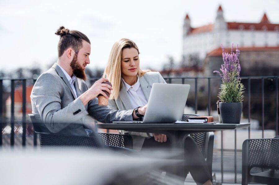 Two cheerful young business people with laptop sitting on a terrace outside office, working on taxes.