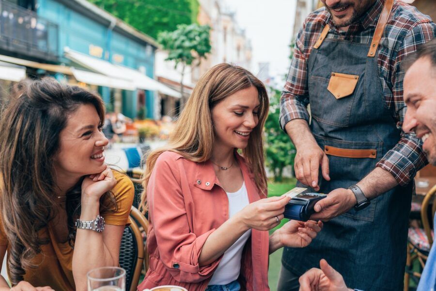 Young woman paying contactless using credit card in sidewalk cafe, with friends.