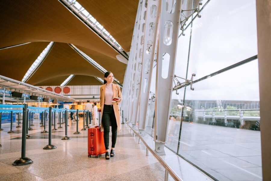 Smiling woman walking toward airport gates, pulling a red suitcase and holding travel documents in her hand