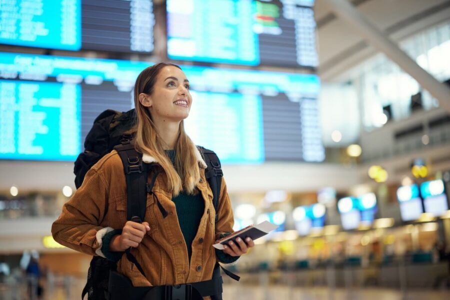 Smiling female backpacker checking her gate on a flight board while holding a passport and boarding pass