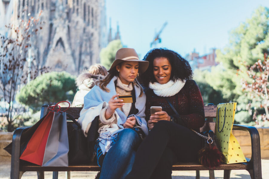 Two friends sitting on a bench using a credit card and a phone