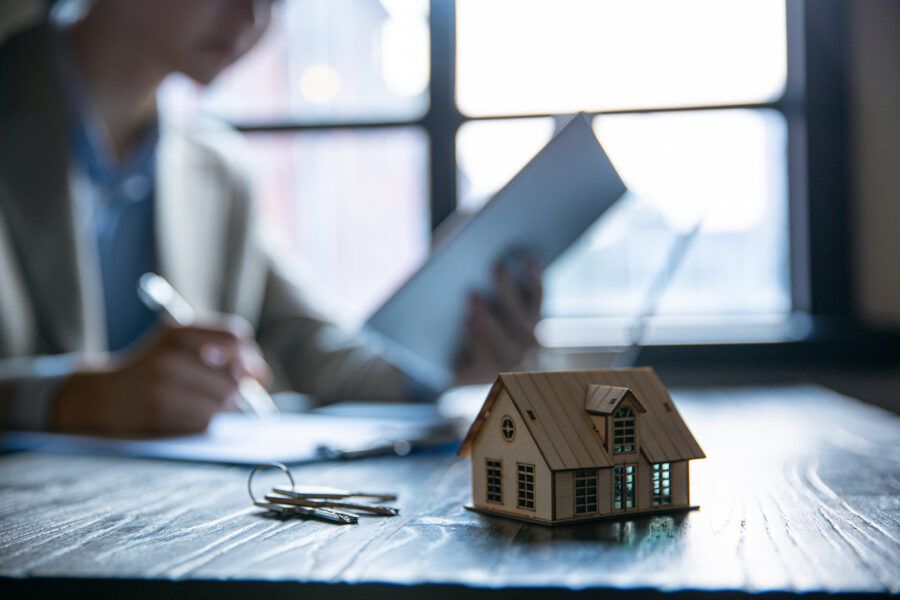 A focused shot of keys and a small model home in the foreground, with a real estate agent looks at paperwork in the background