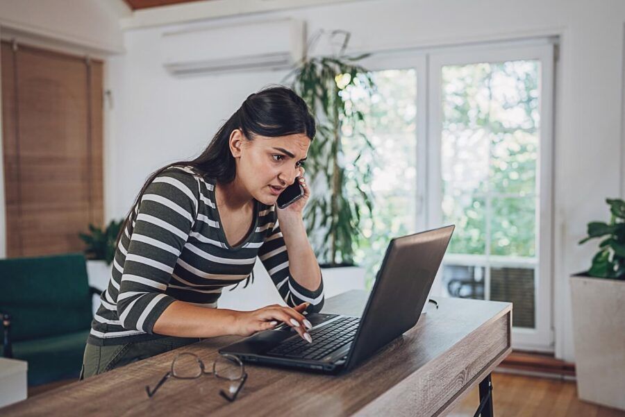 Upset woman standing at a home workstation, holding a smartphone to one ear while using a laptop on a counter. Eyeglasses rest on the desk, and large windows, indoor plants, and modern furnishings are visible in the background