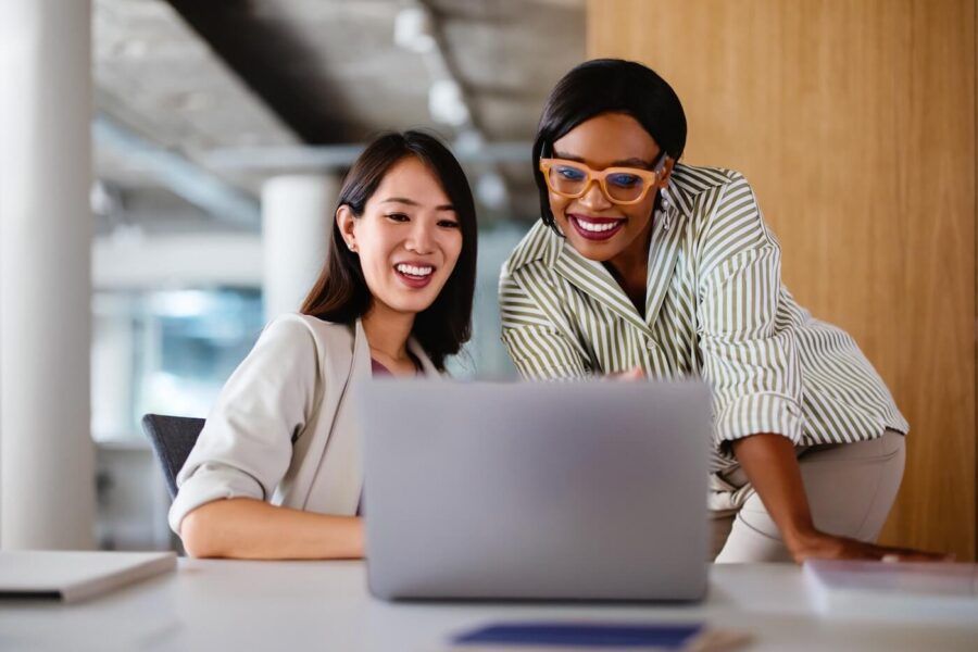 Two smiling female colleagues sitting in an office, using a laptop and discussing something together