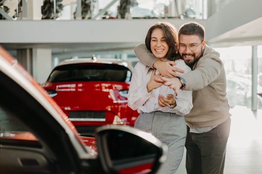 Happy couple shopping for a car; the man hugs the woman with one hand and reaches out to the car with the other hand