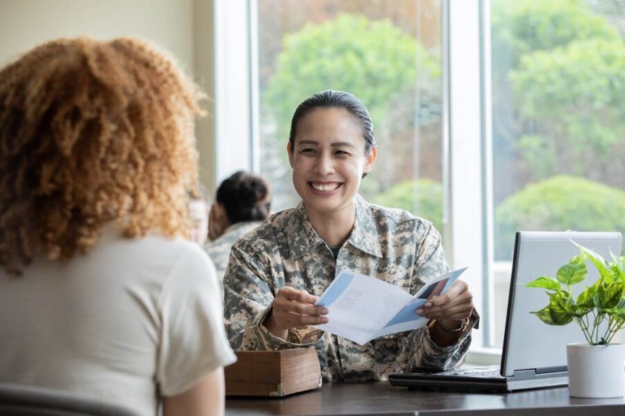 Smiling military woman holding a leaflet and chatting with another woman. A laptop and a potted plant on the desk, other people on the background.