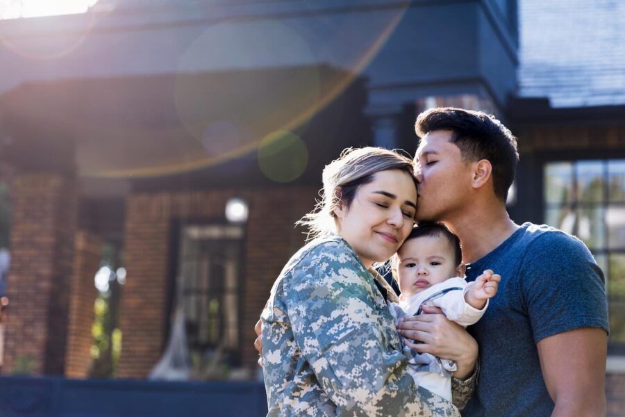 A happy family with a baby standing outside their home; the woman is dressed in a military uniform
