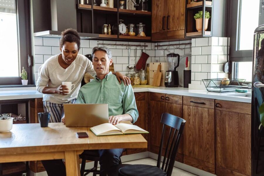 Mature couple at a kitchen table reviewing a laptop together, with an open notebook, phone, and coffee mug nearby.