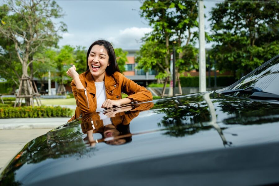 Young woman resting on the hood of her car, celebrating saving money on car expenses.