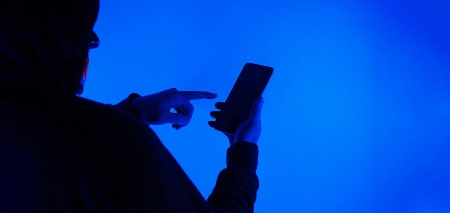 Anonymous hacker with smartphone in hand. His hooded silhouette is against a blue background.