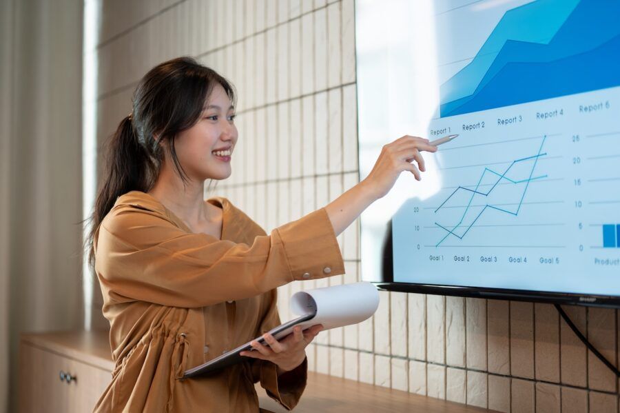 A woman is pointing a pen at the monitor and presenting her investing information.