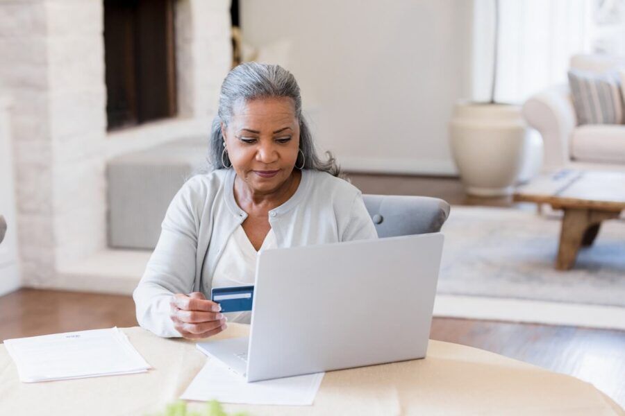 Senior woman sitting at a dining table, using a laptop while holding a credit card, with paperwork spread out in a bright living room