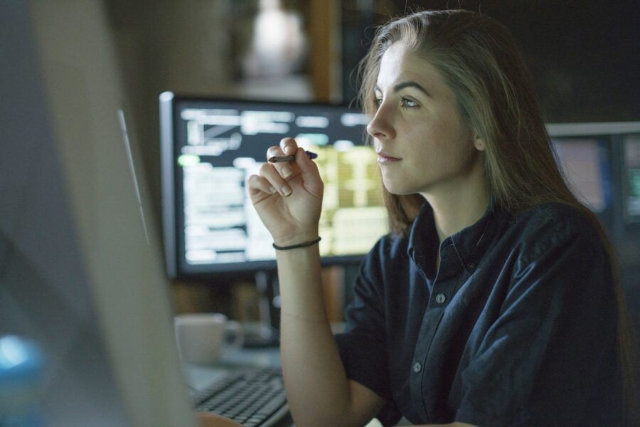 A young woman is seated at a desk surrounded by monitors, contemplating work in her office.