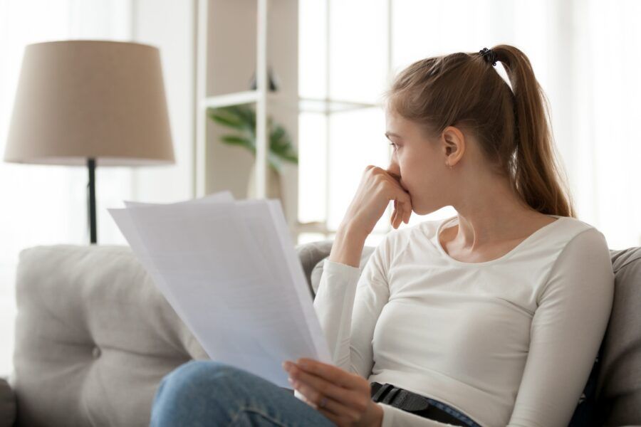 Upset thoughtful woman holding paper document in hands, sitting on sofa, looking in distance.