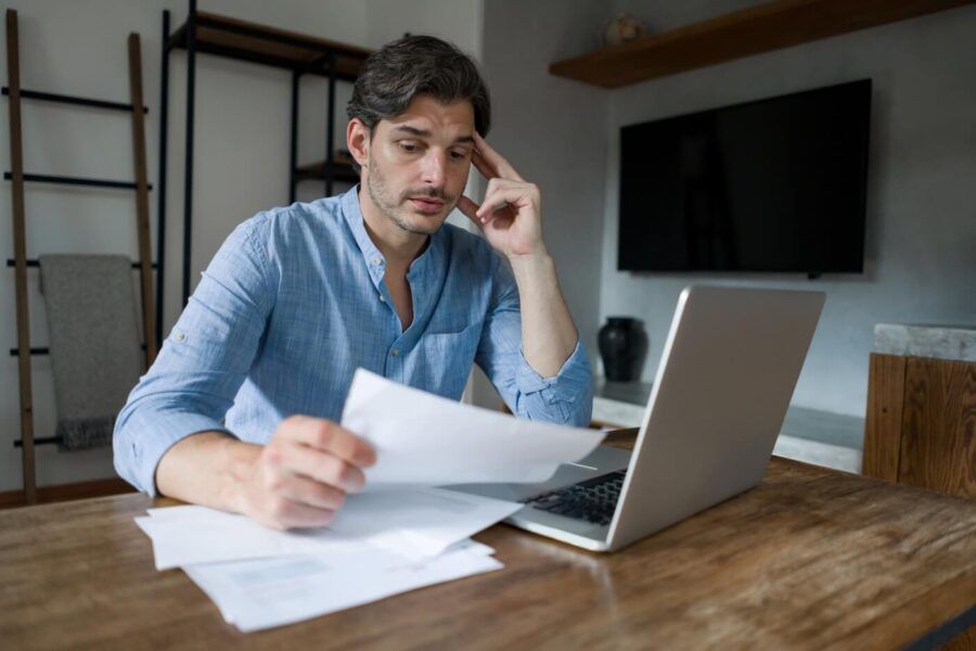 Frustrated young man examining paperwork in his home office, with an open laptop on the desk beside him