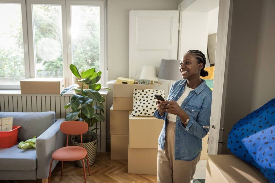 Smiling woman standing in a room filled with moving boxes, holding a smartphone near the doorway.