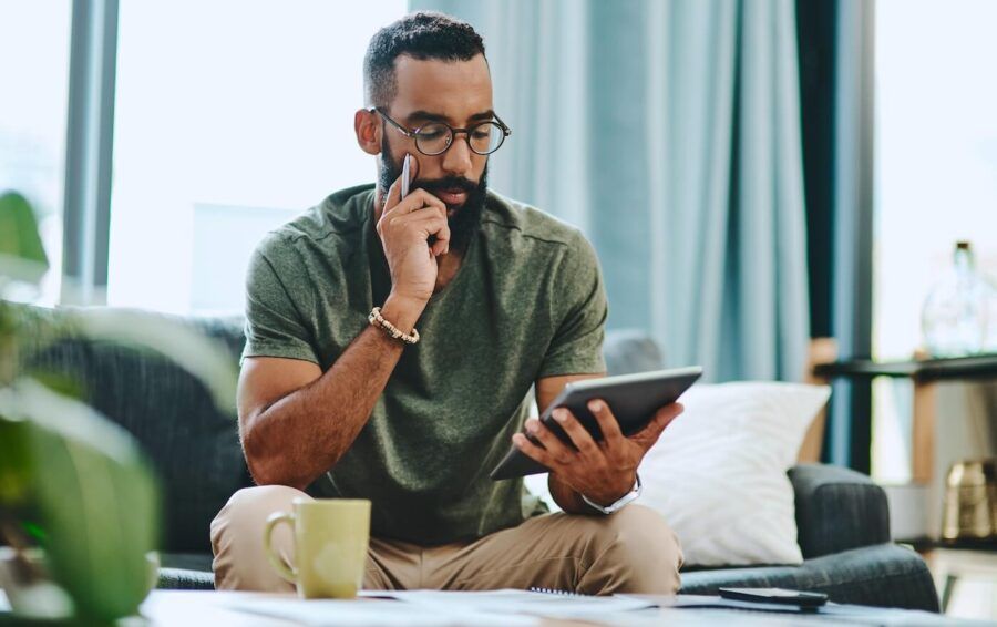 Focused man sits in a living room using a tablet while reviewing paperwork