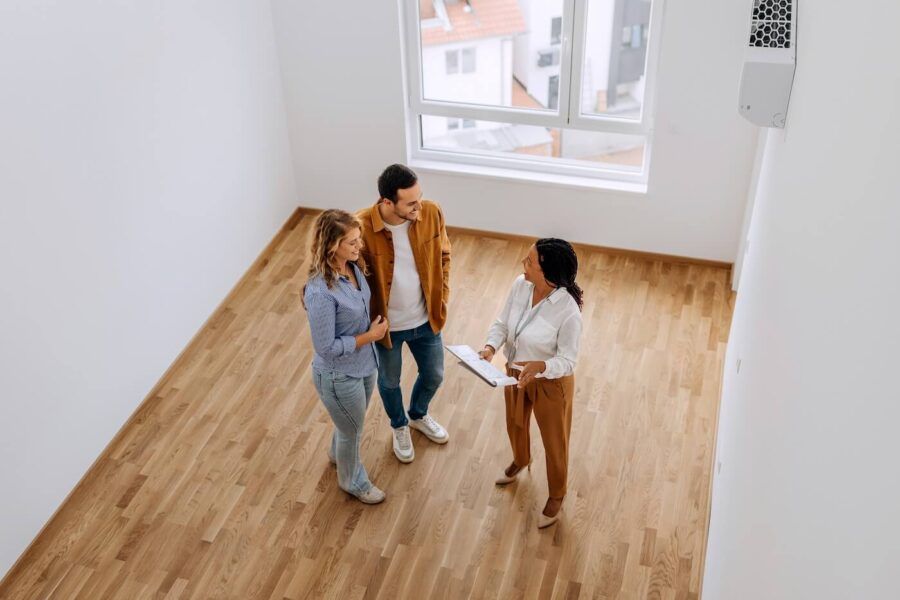 Photo of a couple and a female real estate agent chatting in an empty room with white walls and hardwood floor, taken from above