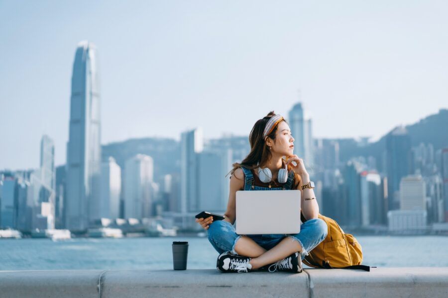 A young woman sitting cross-legged by the promenade, against urban city skyline. She is wearing headphones around neck, using smartphone and working on laptop, with a coffee cup by her side. Looking away in thought.