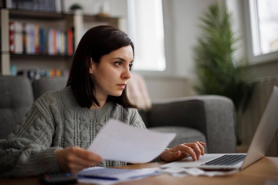Focused young woman holding a printout while looking at the laptop screen