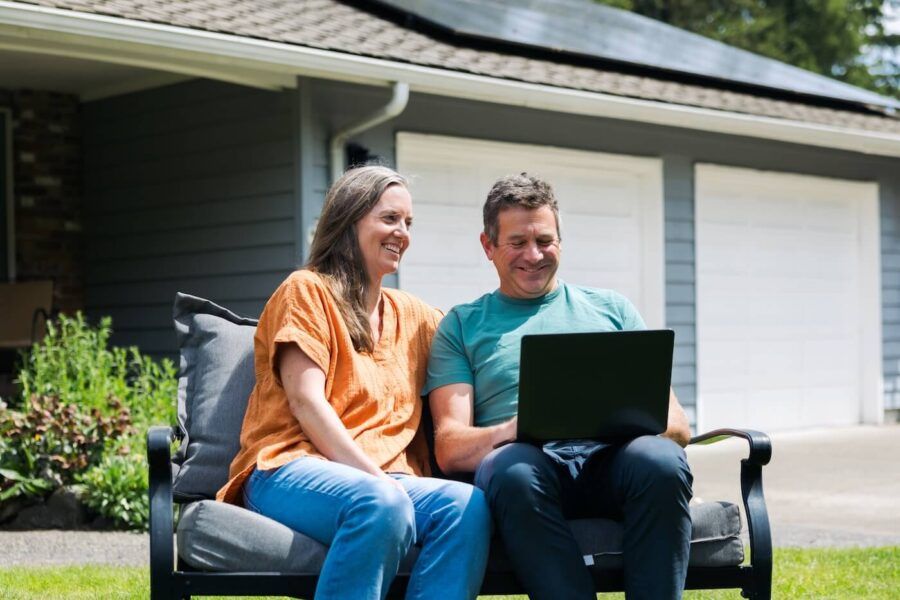 Mature couple sitting together on an outdoor patio chair in front of a house with an open laptop, with a garage door, siding, greenery, and lawn visible in the background.