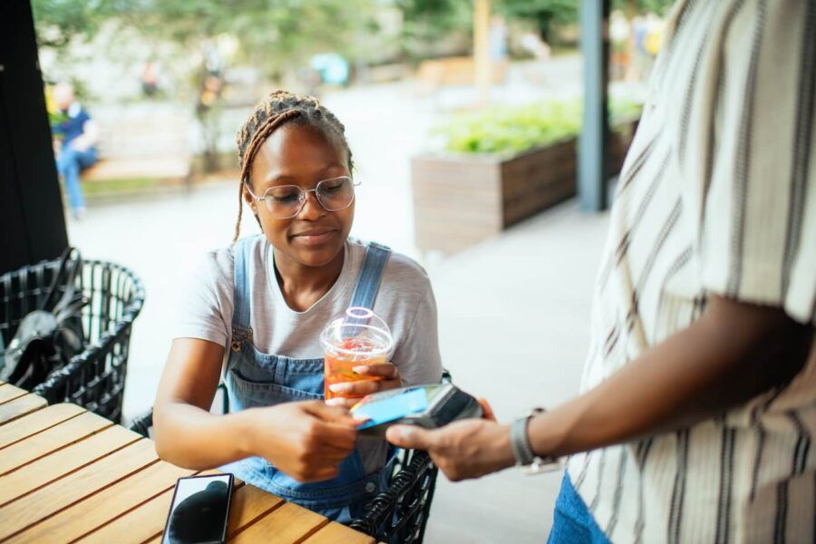 Young woman wearing glasses paying with credit card in the outdoor cafe while holding a beverage