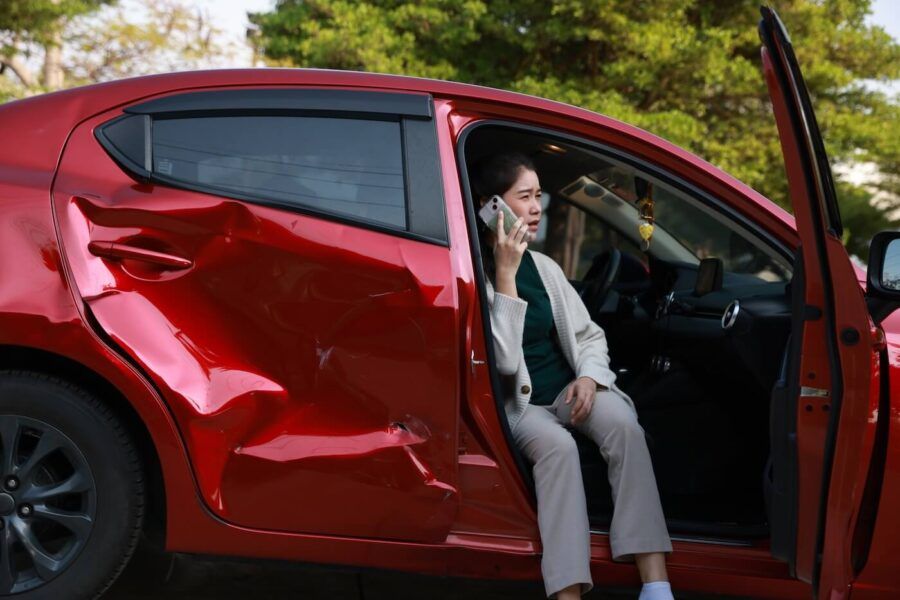 Upset woman sitting in a damaged red car on a passenger seat and making a call to the insurance company