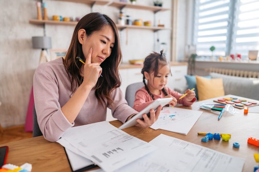 Focused young woman reviewing printed documents while a toddler girl playing beside her.