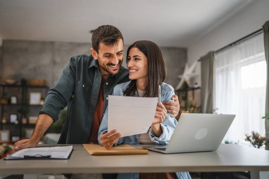 Happy couple standing close together at a table while looking at a document, with an open laptop, notebook, and papers arranged on the surface in a home setting.