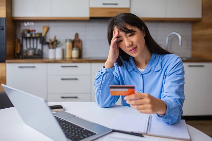 Worried woman holding a credit card while sitting at the kitchen table, with a laptop and a notepad beside her