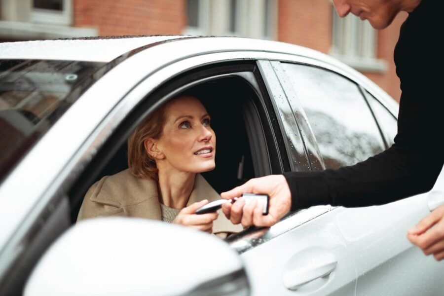 A woman sits inside a parked car while handing keys to another person standing outside the vehicle