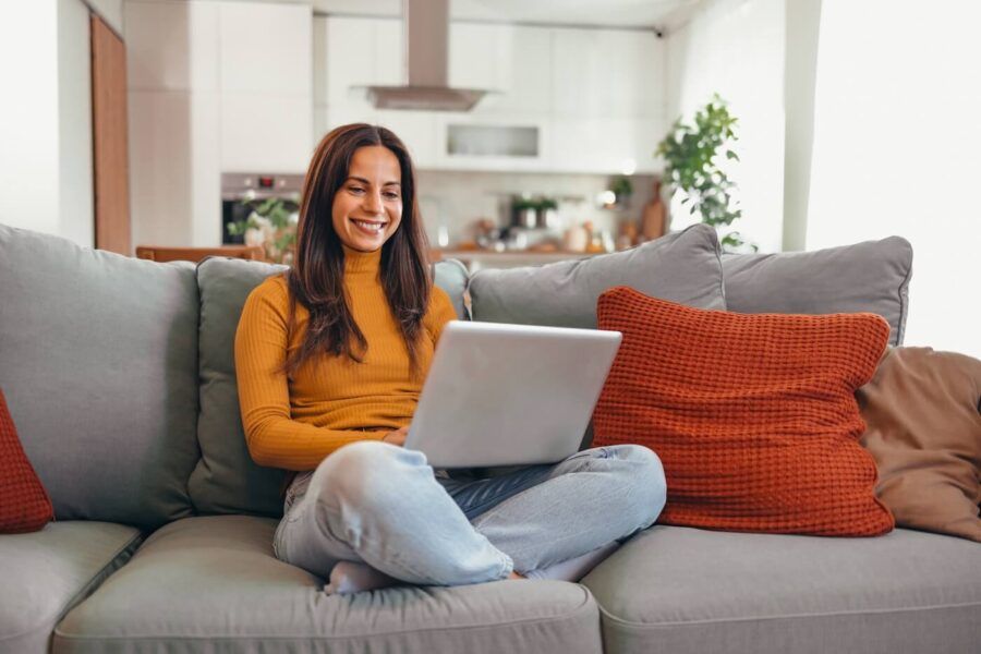 Smiling young woman sitting on the sofa with crossed legs and using her laptop