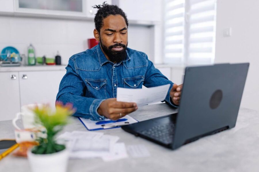 Focused man sits at a table holding papers while looking at a laptop, with additional documents and a pen on the table