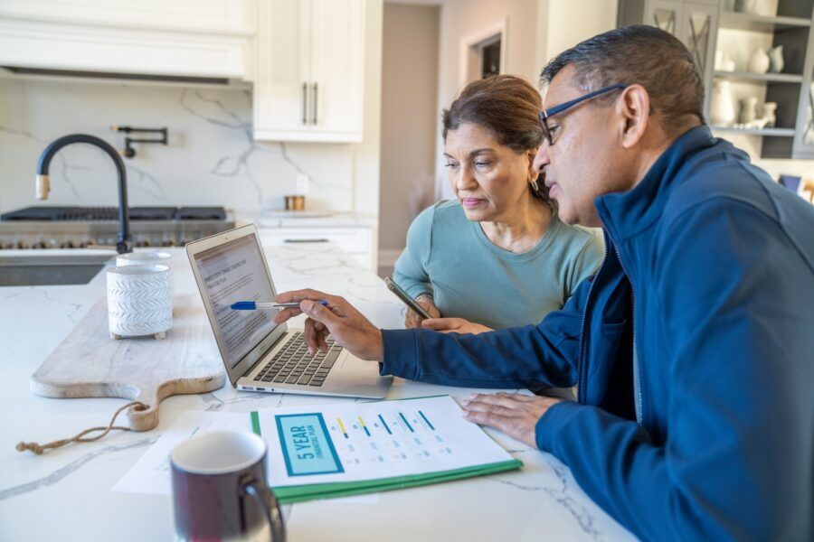 A couple sit at their kitchen island with a laptop open and their bills spread out in front of them as they review their finances together. They are dressed casually and appear inquisitive.