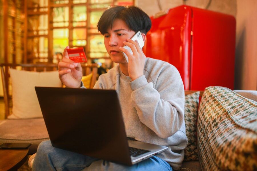 A young man sitting on sofa and looking at credit card and laptop
