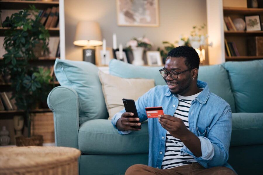 Smiling man wearing glasses sitting on the floor next to a couch and using his mobile phone and a credit card