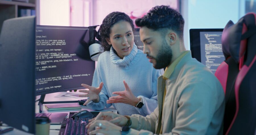 A woman coaching a man on cybersecurity essentials. They are sitting at desks with many computer monitors showing code.